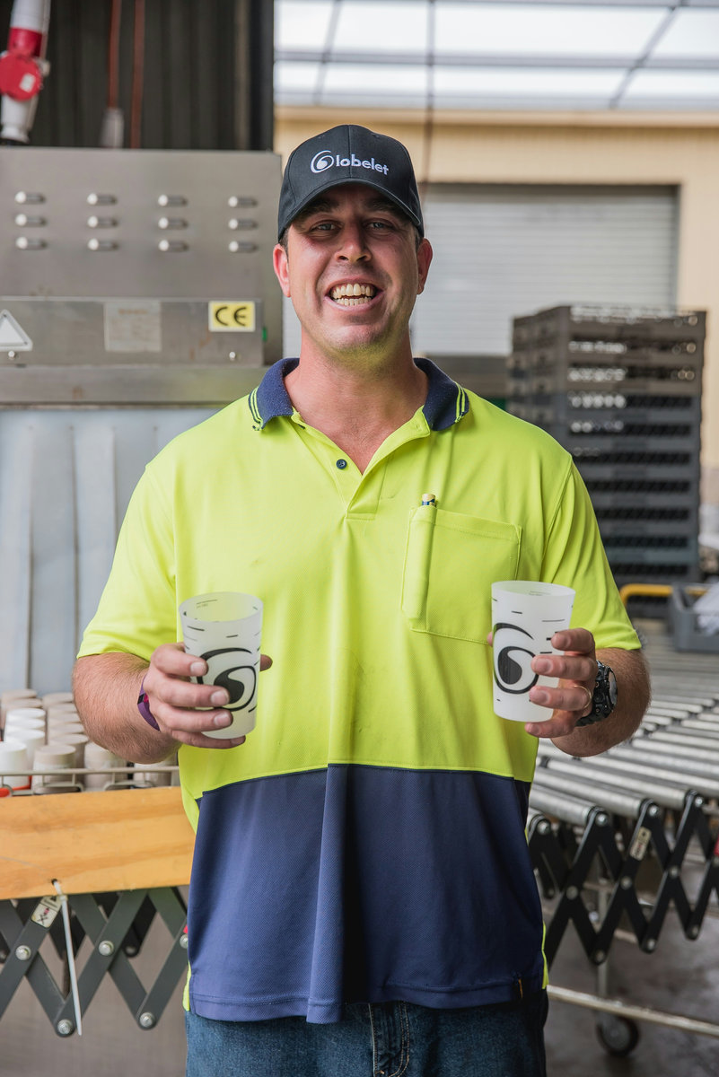 A warehouse worker in a hi-vis polo shirt, smiling at the camera and holding two cups.